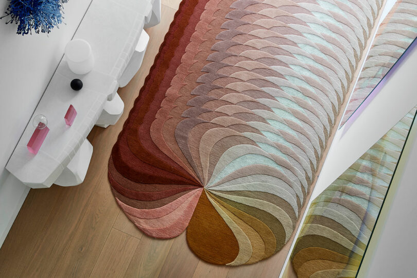 A multicolored, fan-shaped rug with overlapping arcs sits on a wooden floor next to a white console table with decorative objects.