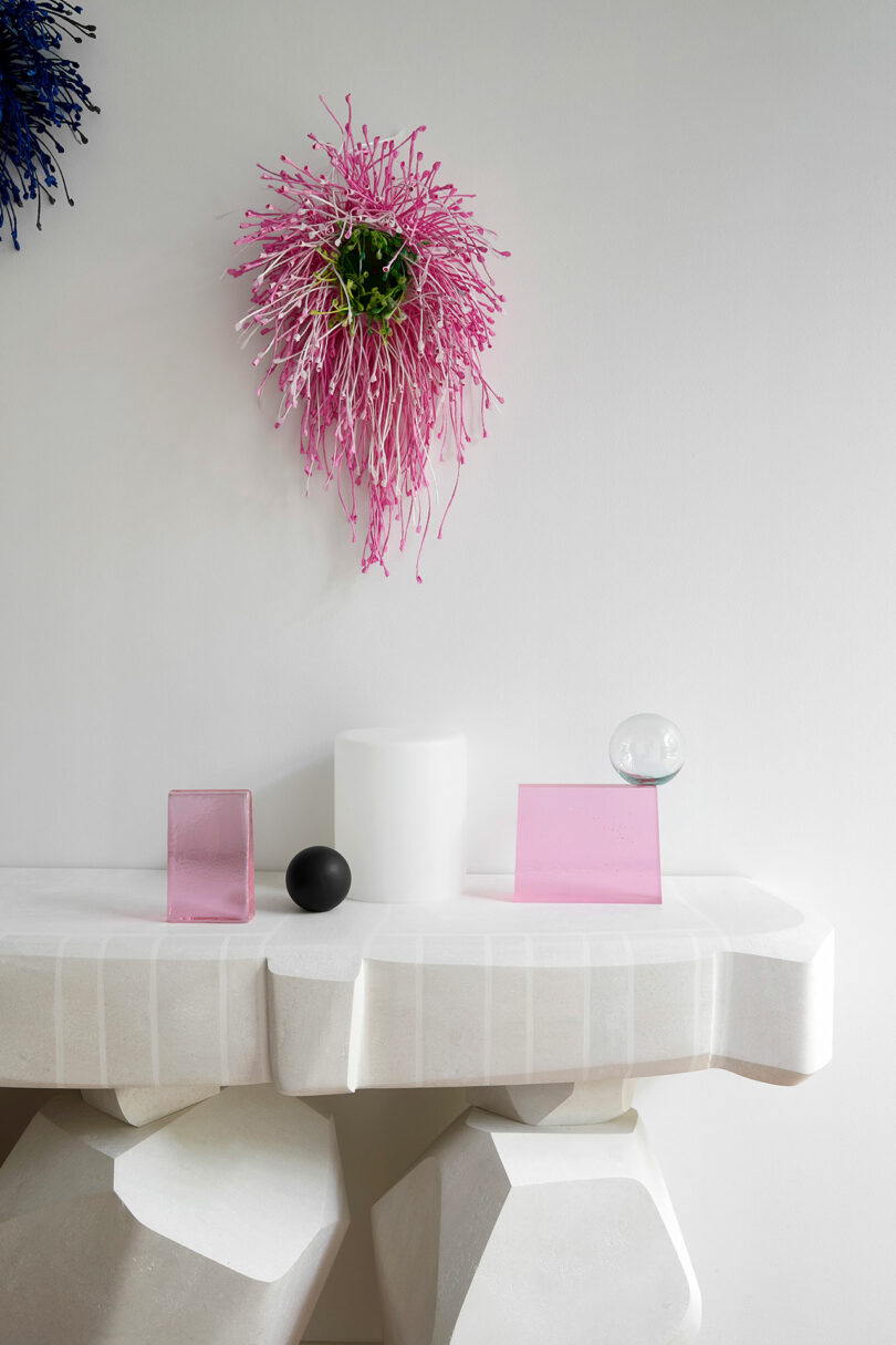 A white geometric table with pink glass blocks, a black sphere, a clear sphere, a white cylinder, and a pink wall-mounted abstract floral sculpture above it.