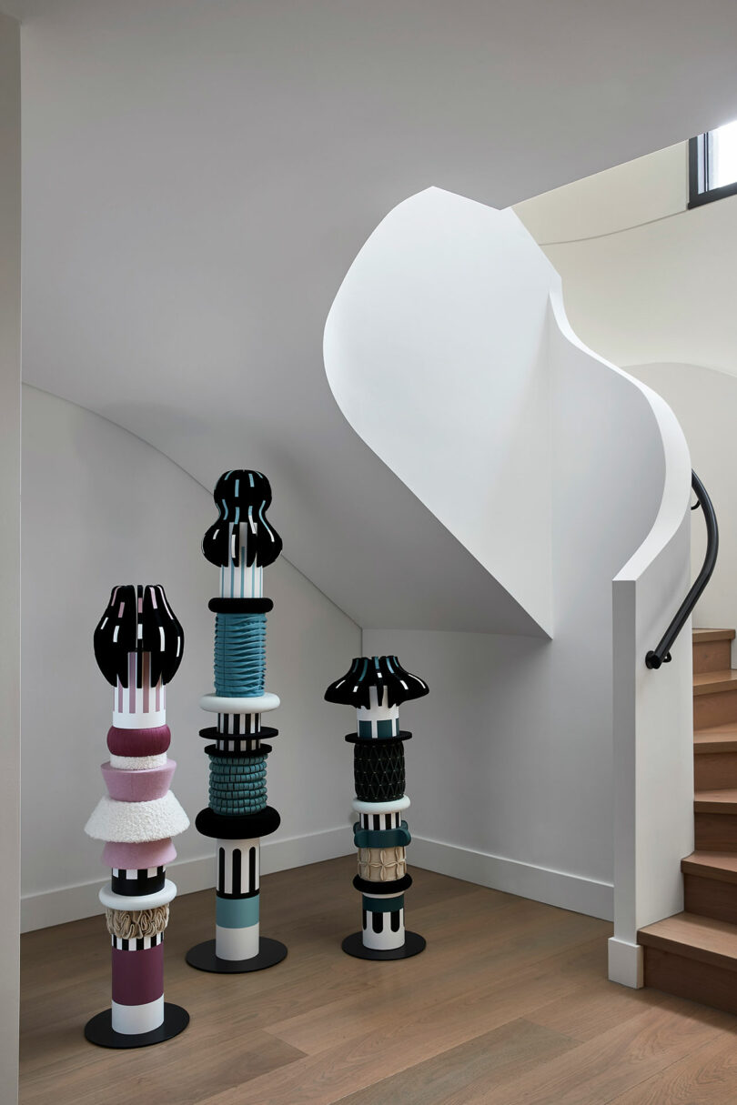 Three colorful, abstract sculptural columns stand beneath a modern white staircase with wooden steps and a curved handrail in a minimalist interior.