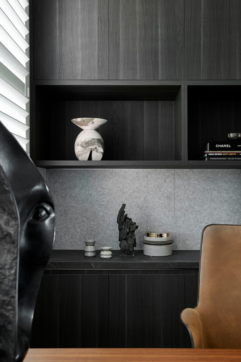 Modern office with dark wood shelves, decorative vases, sculptures, books, and a tan leather chair; partial view of an abstract black sculpture in the foreground.