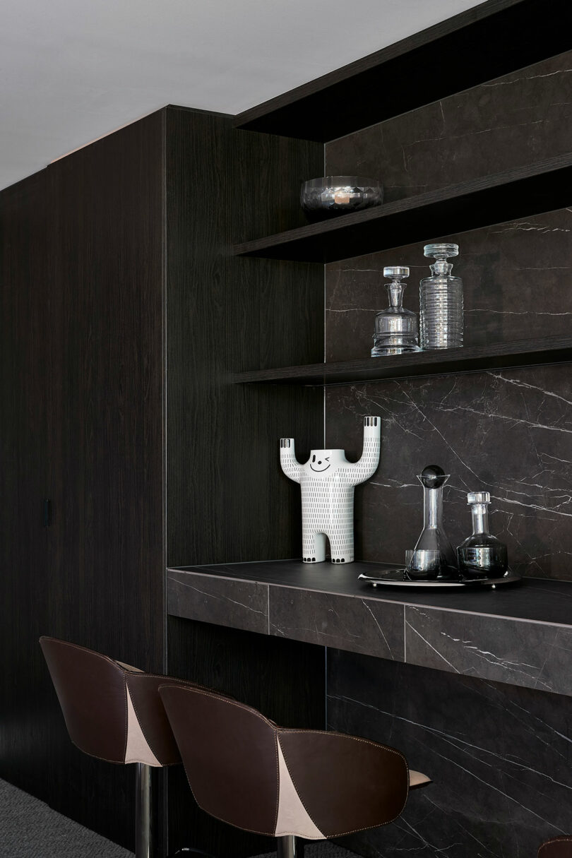 A modern bar area with dark wood paneling, glass decanters, a white decorative figure, and two brown bar stools.