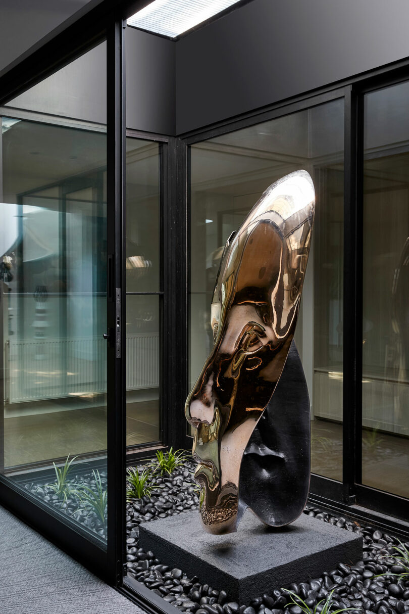 A polished, elongated metallic sculpture of a human face is displayed on a stone base inside a glass-walled room with black pebbles and small plants.