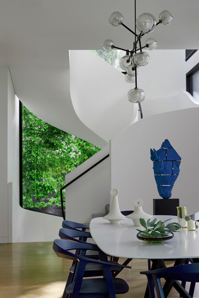 Modern dining area with a white table, blue chairs, abstract sculptures, a black chandelier, large window with greenery, and a curved white staircase in the background.