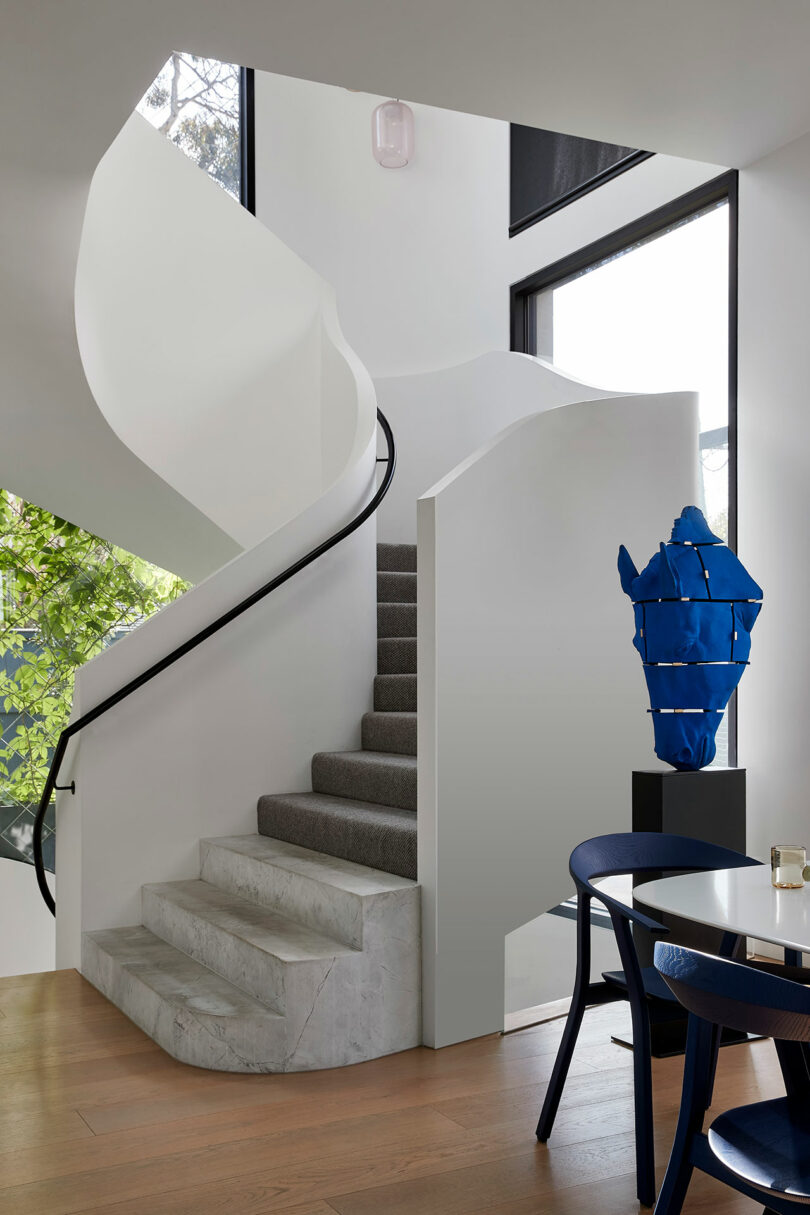 A modern interior with a curved white staircase, marble steps, black handrail, large window, wooden floor, blue abstract sculpture, and navy chairs around a round table.