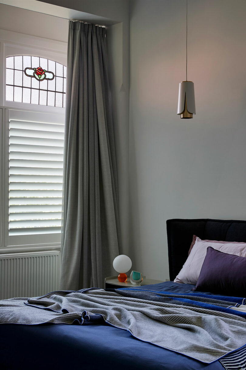 A modern bedroom with a blue bed, purple pillows, bedside table with a lamp, grey curtains, white shutters, and a stained glass window above.