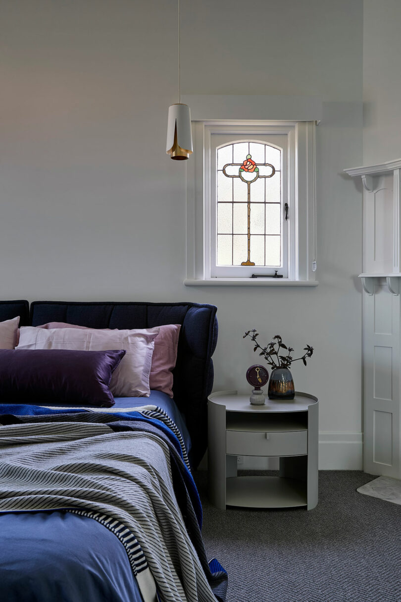 A neatly made bed with purple and blue linens sits beside a small round nightstand, under a decorative stained glass window in a softly lit bedroom.