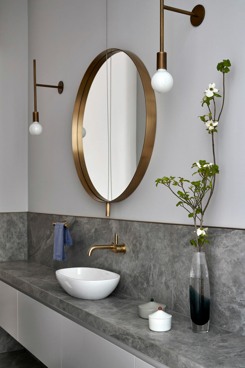 Modern bathroom with a round brass-framed mirror, wall-mounted lights, marble countertop, vessel sink, and a vase with branches on the right.