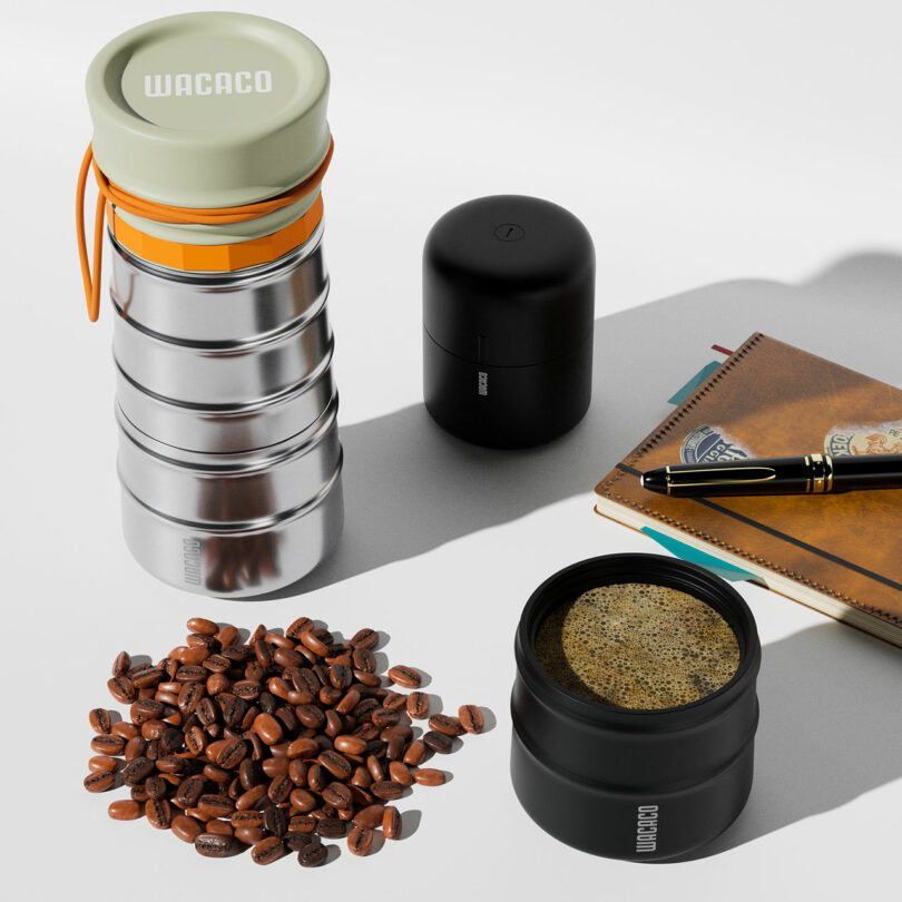 A portable coffee maker, a black canister, notebook with pen, loose coffee beans, and a cup of brewed coffee are arranged on a white surface.