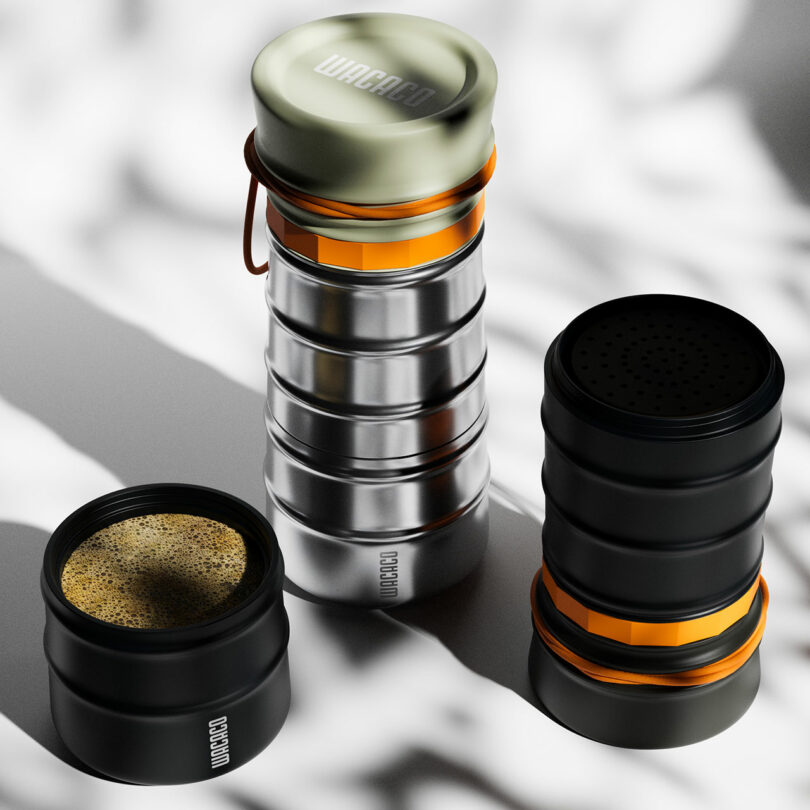Three stainless steel and black coffee brewing containers with orange accents are shown; one is open with coffee inside, and two are closed, set on a light, shadowed surface.