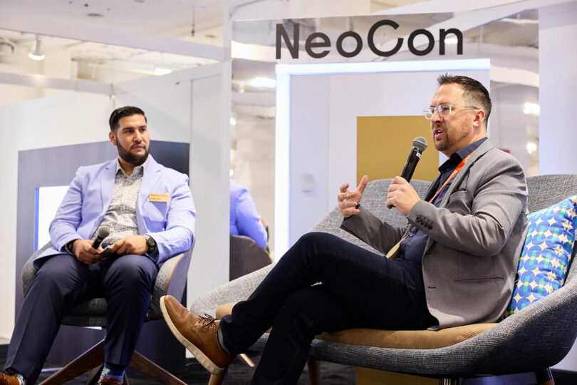 Two men sit and talk at a NeoCon panel discussion; one holds a microphone while the other listens attentively.