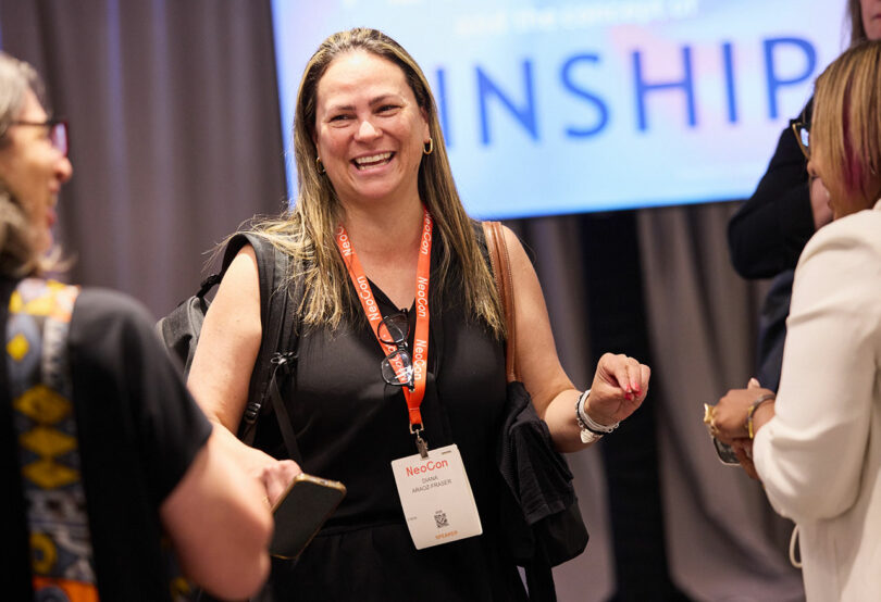 A woman wearing a NeoCon badge and orange lanyard smiles while conversing with others at an indoor event.
