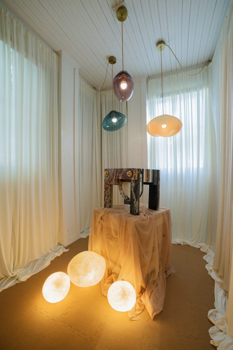 A small room with sheer white curtains, three glowing orbs on the floor, a draped table, and three pendant lamps hanging from the ceiling