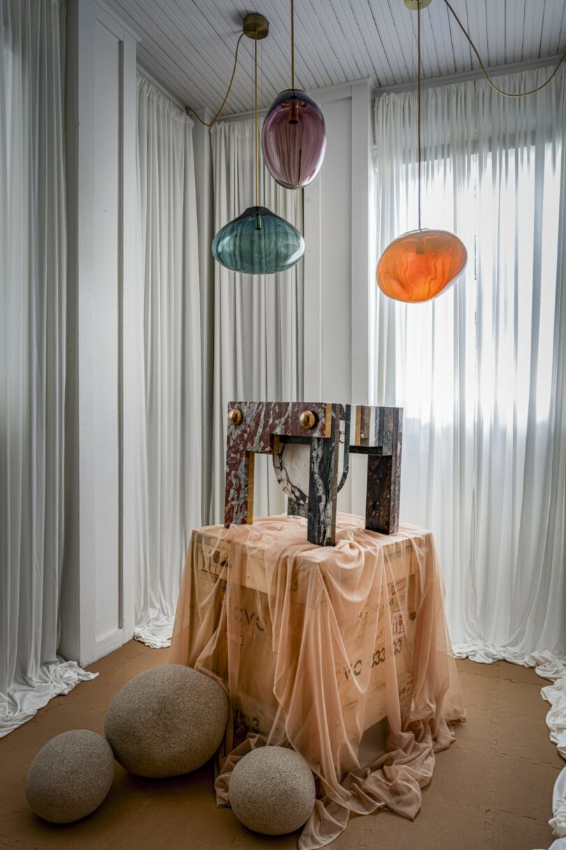 A decorative room corner with sheer white curtains, three colorful pendant lights, a marble console on a draped box, and several round stone-like lamps on the floor