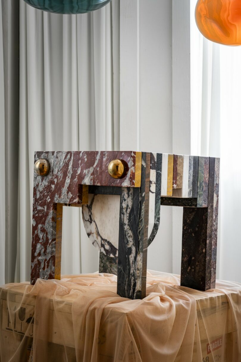 A colorful marble table with geometric shapes and two round gold knobs stands on a draped crate in a room with white panel walls and hanging glass lamps above
