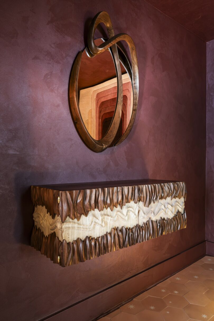 A sculptural wooden floating cabinet with wavy textured details is mounted below an abstract round wooden mirror on a maroon wall