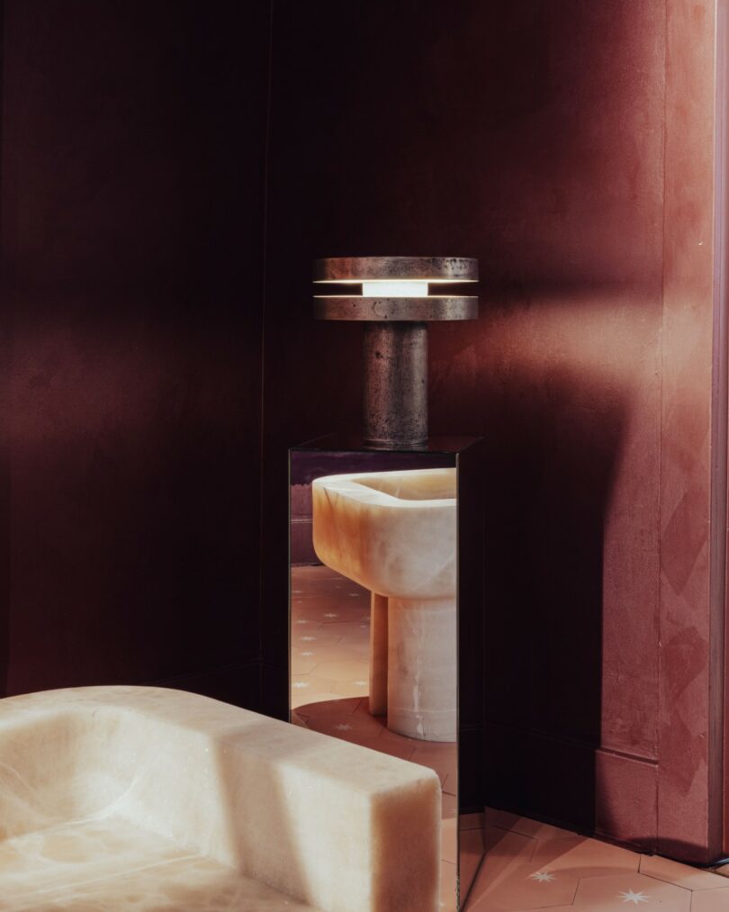 A modern marble or stone sink is reflected in a mirror beside a cylindrical lamp, set in a room with dark red walls and soft lighting
