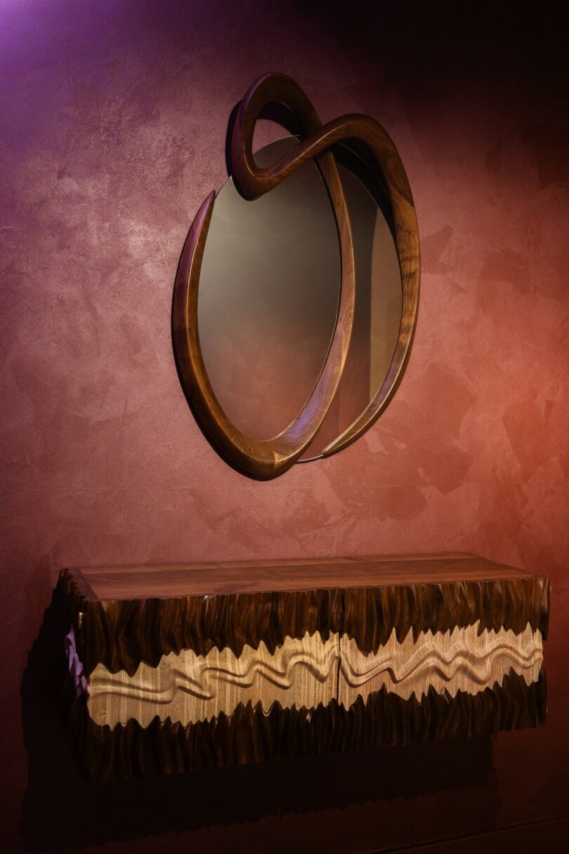 A decorative mirror with a curved wooden frame hangs above a wooden console table with a wavy, layered edge against a textured maroon wall