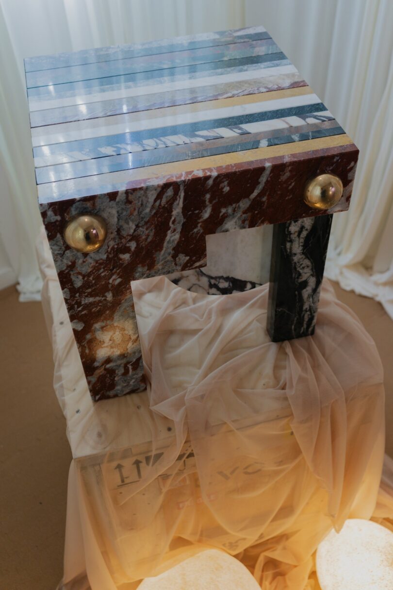 A geometric table with a multicolored, striped stone surface and gold sphere accents stands draped in translucent fabric on a wooden crate, surrounded by soft lighting