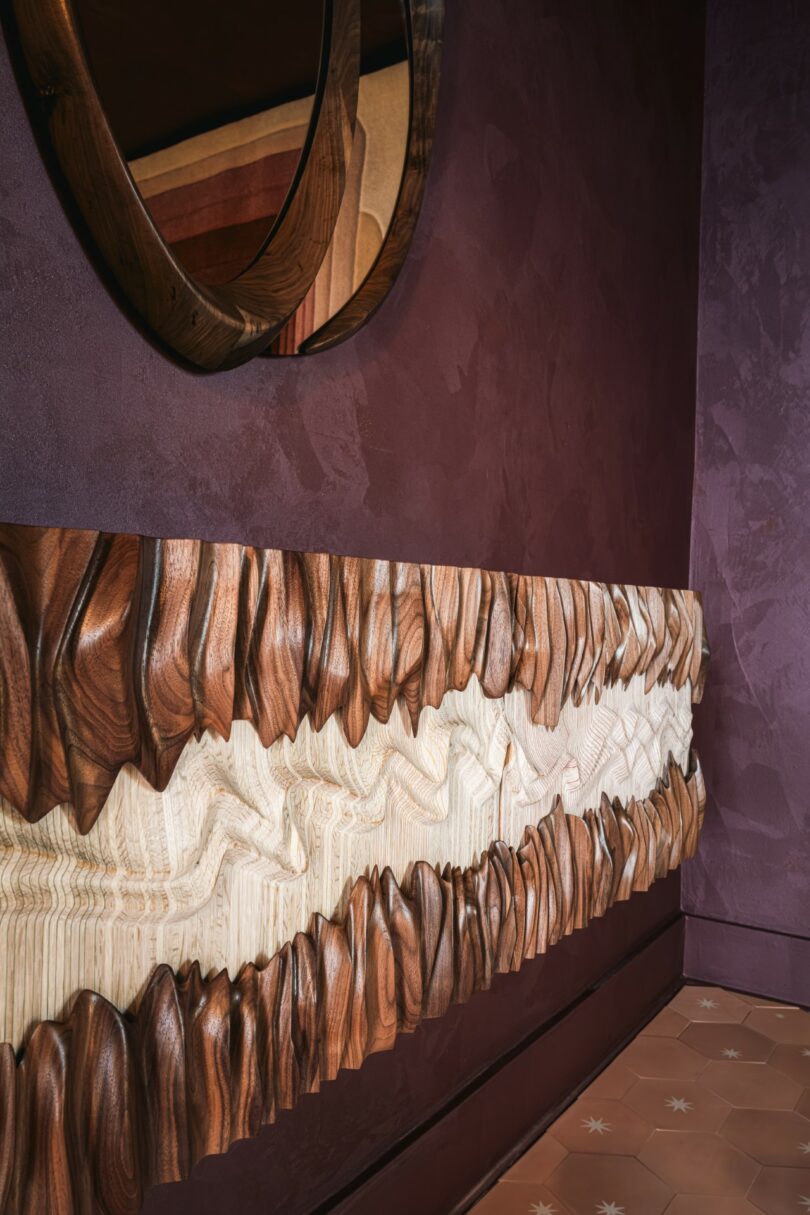 A textured wooden wall panel with alternating dark and light carved waves, set against a maroon wall beneath a round mirror