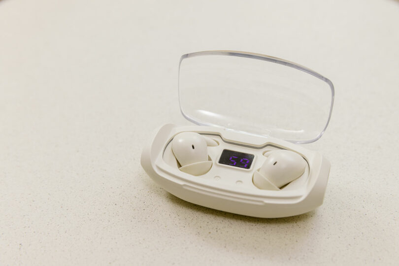 A pair of white SleePods wireless earbuds in a charging case with a transparent lid and digital display, placed on a light-colored surface.