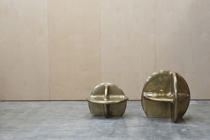 Two shiny, sculptural brass chairs of different sizes are placed side by side on a concrete floor against a plain wooden wall.