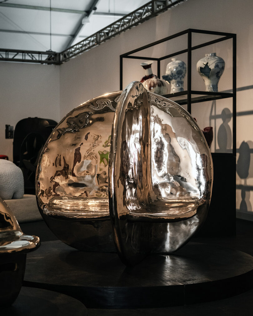A shiny, metallic spherical sculpture with a split design is displayed on a pedestal in an art gallery with ceramics on shelves behind it.