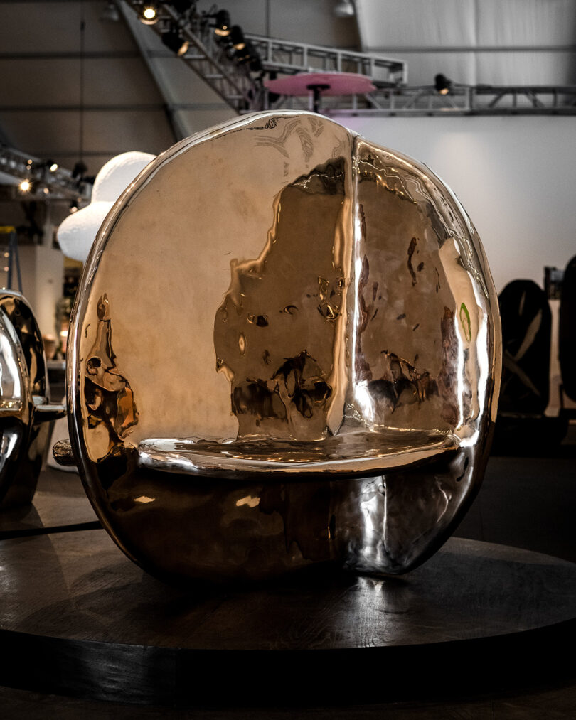 A polished metallic sculptural bench with a rounded, reflective surface displayed in an indoor exhibition space.