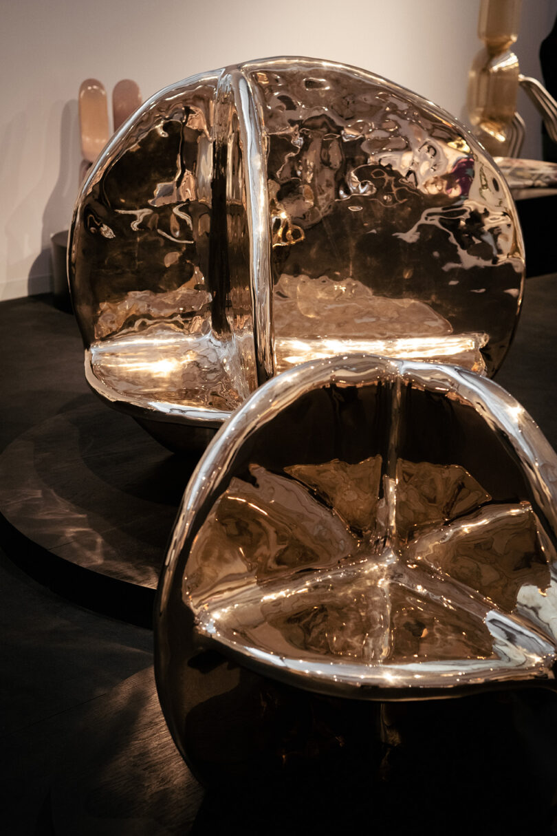 Two polished, metallic sculptural chairs with organic, rounded forms are displayed on a dark floor in an indoor setting.