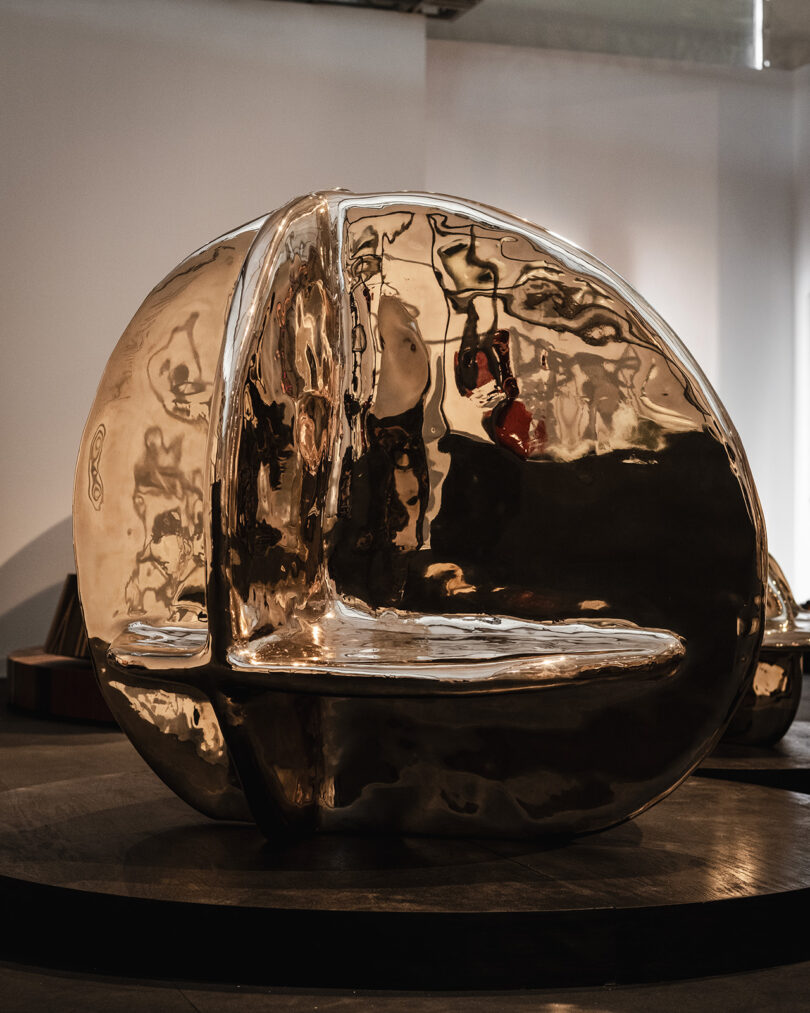 A large, reflective, golden abstract sculpture with smooth, curved surfaces is displayed indoors on a dark circular platform.