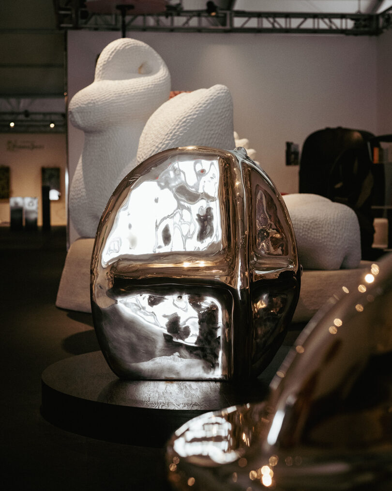A reflective metallic abstract sculpture is displayed on a round pedestal, with large textured white sculptures in the background within an indoor exhibition space.