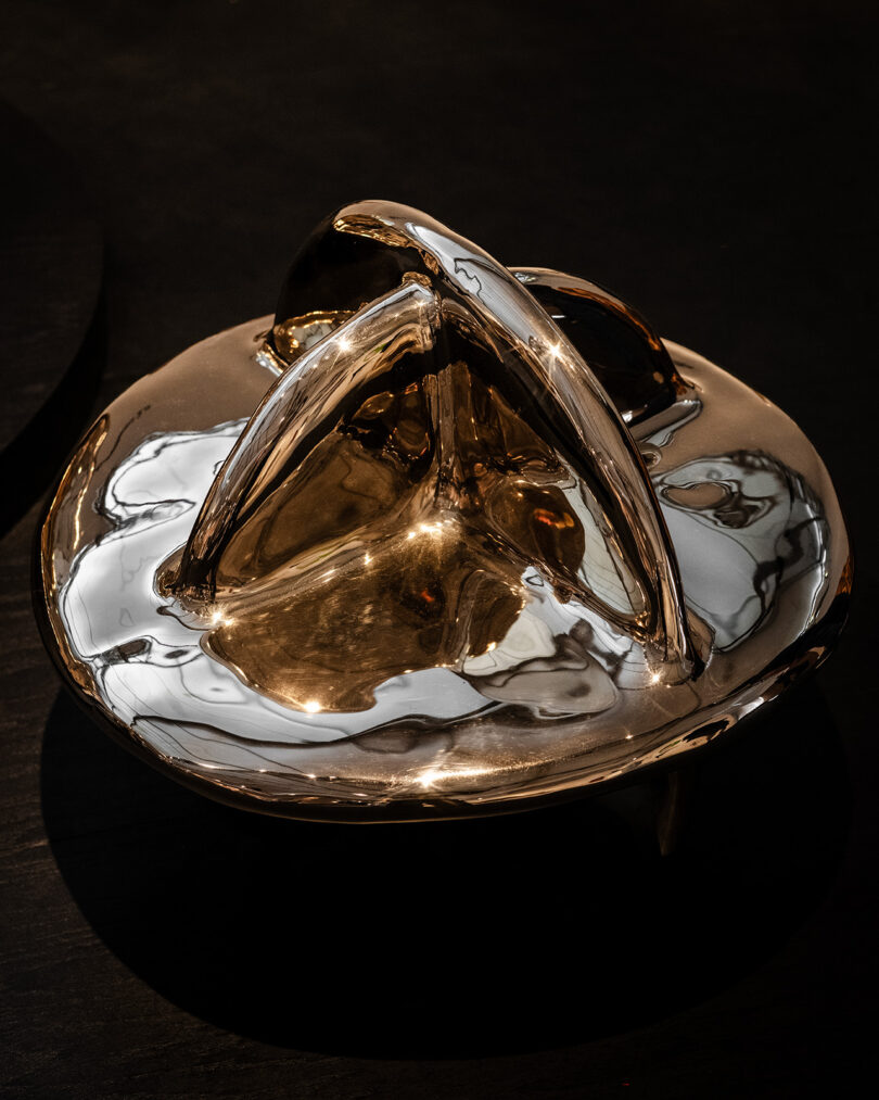 A polished, abstract metallic sculpture with curved, intersecting arches is displayed on a dark surface.