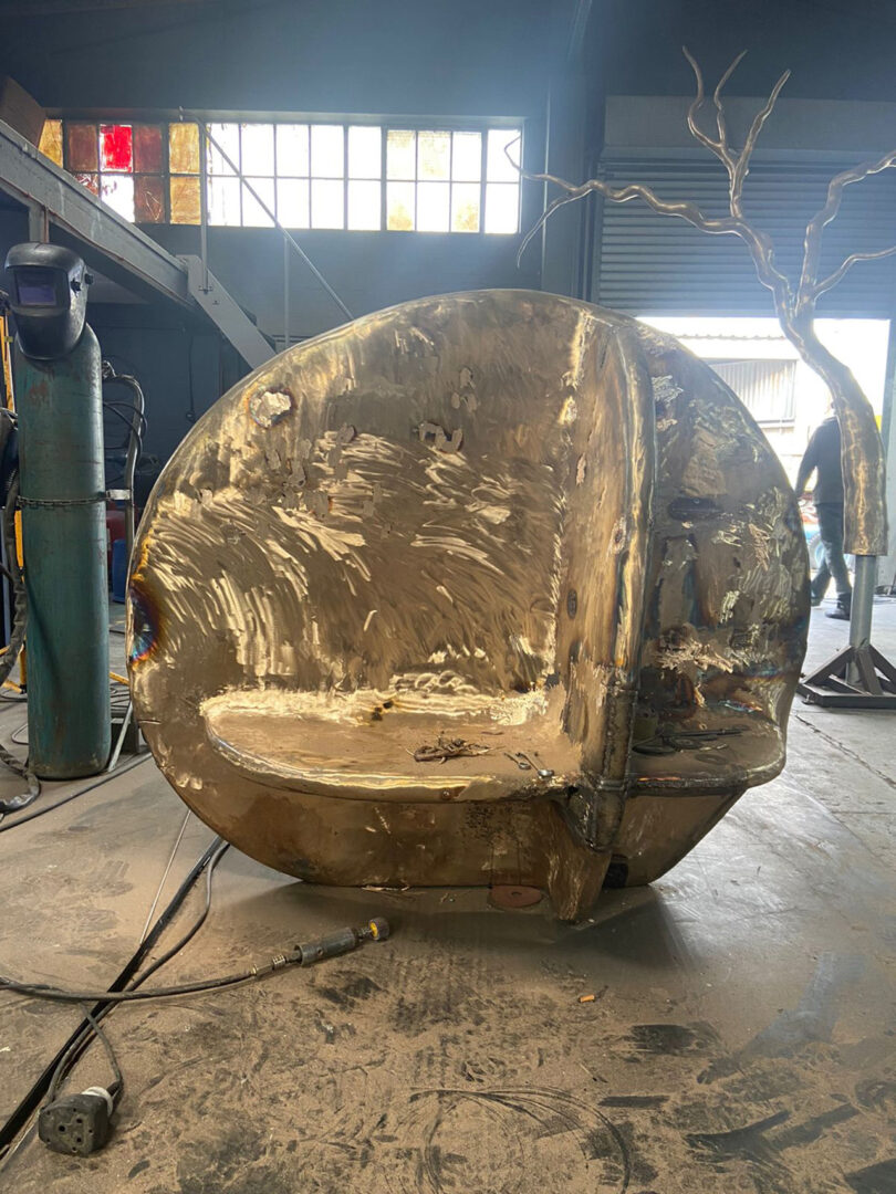 A large, unfinished metallic sculpture shaped like a circular bench sits in an industrial workshop with tools and welding equipment nearby.