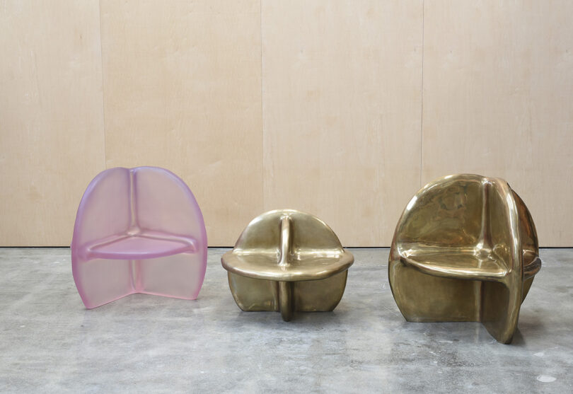 Three sculptural chairs are arranged side by side: one translucent pink and two metallic gold, set against a light wooden wall and concrete floor.