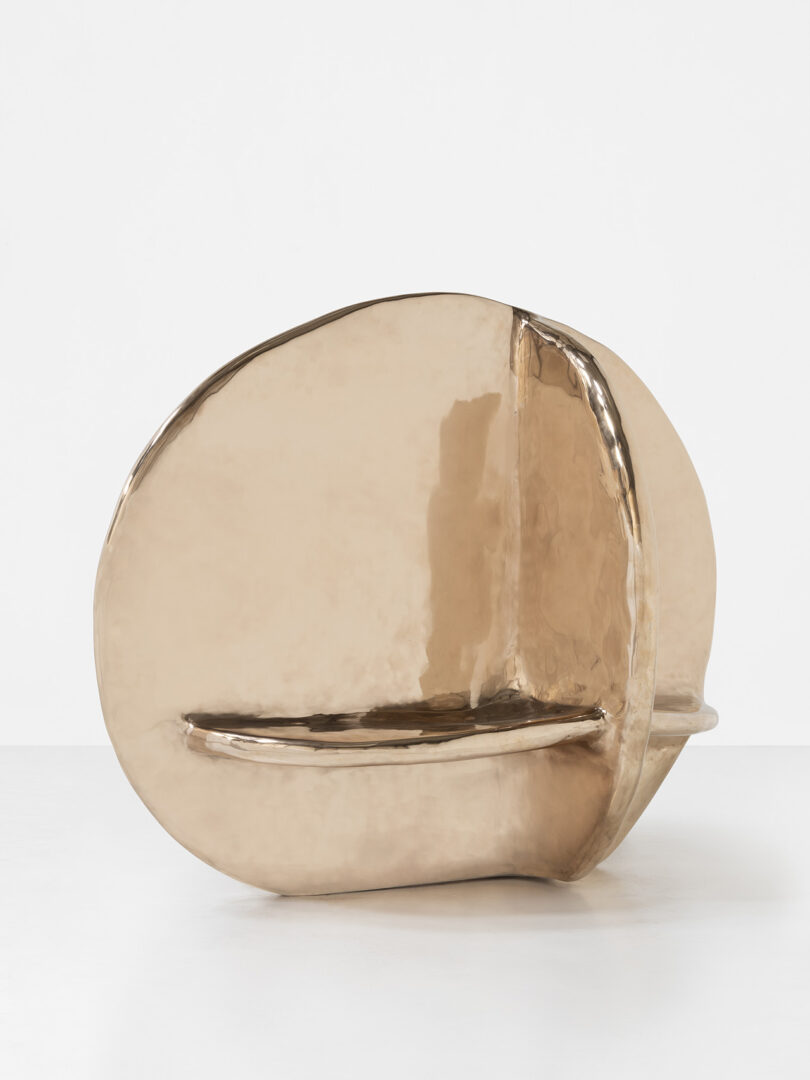 A sculptural, metallic, bronze-colored bench with a rounded, abstract shape and a smooth, reflective surface, set against a plain white background.