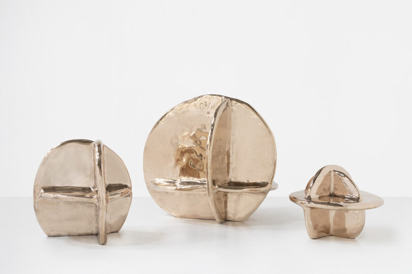 Three abstract, metallic bronze sculptures of varying sizes with intersecting curved and straight planes, displayed on a plain white surface against a white background.