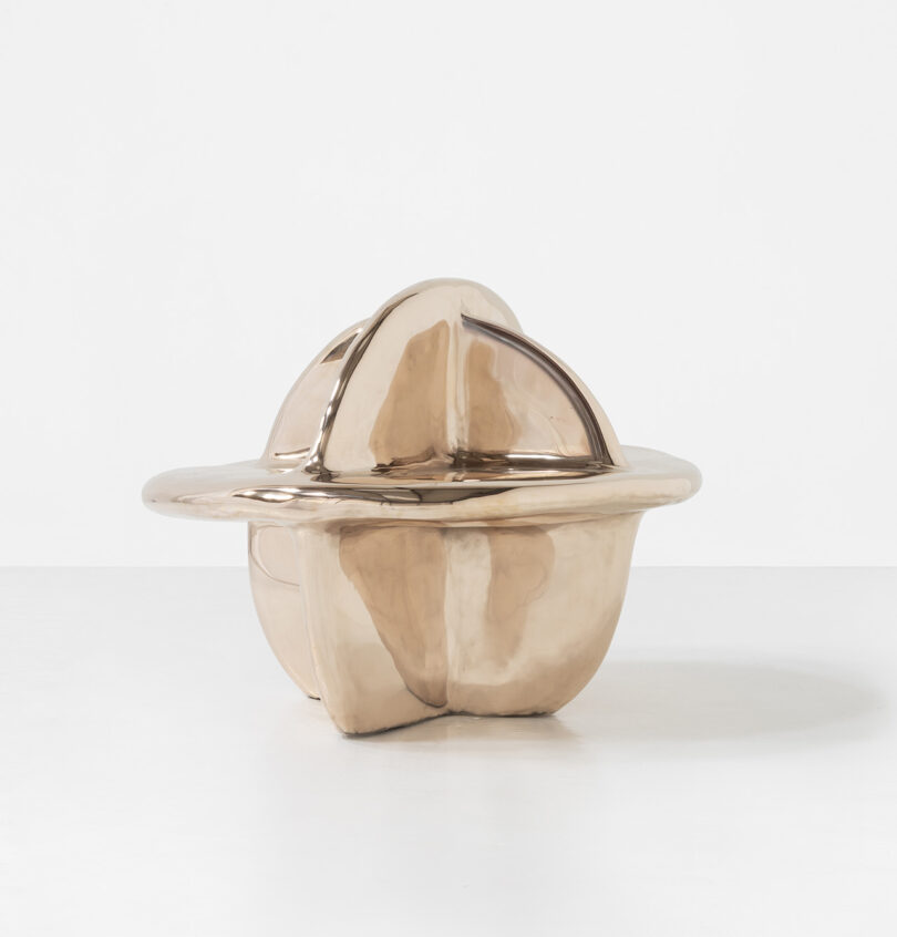 A smooth, metallic, bronze-colored sculptural object with rounded curves and intersecting dome shapes, set against a plain white background.