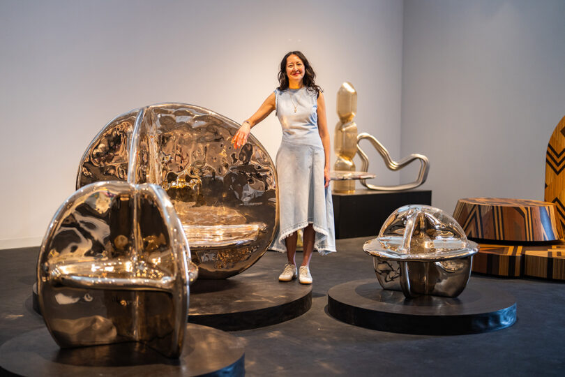 A woman in a light dress stands smiling beside large, reflective, metallic sculptural furniture pieces in an art gallery.