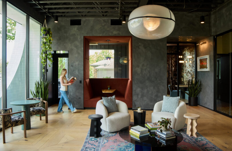 Modern lounge with armchairs, tables, books, plants, and a large Powell overhead light; a person walks by expansive windows, and a built-in seating alcove is visible in the background.