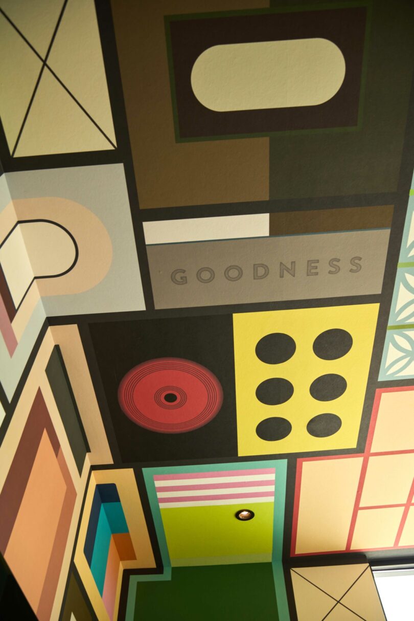 Ceiling mural with geometric shapes, circles, and rectangles in various colors; the word “GOODNESS” is printed in uppercase letters among the designs, reflecting Powell’s vibrant artistic style.