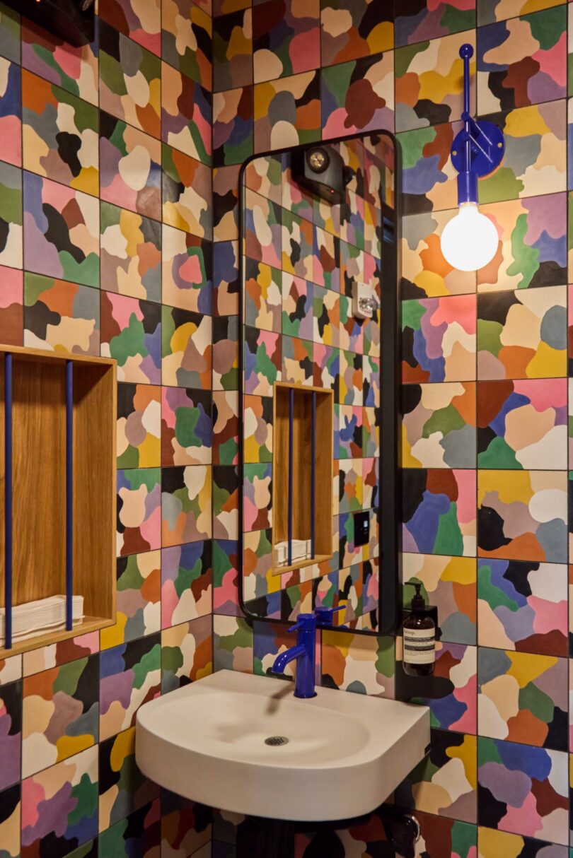 A small Powell bathroom with multicolored camouflage-patterned tiles, a rectangular mirror, a white sink, blue faucet and light fixture, and a round exposed bulb.