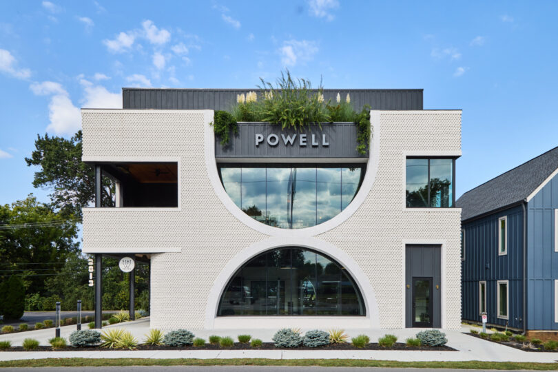 Modern three-story Powell building with a geometric white facade, large arched window, prominent "POWELL" sign at the center, and lush greenery adorning the rooftop.