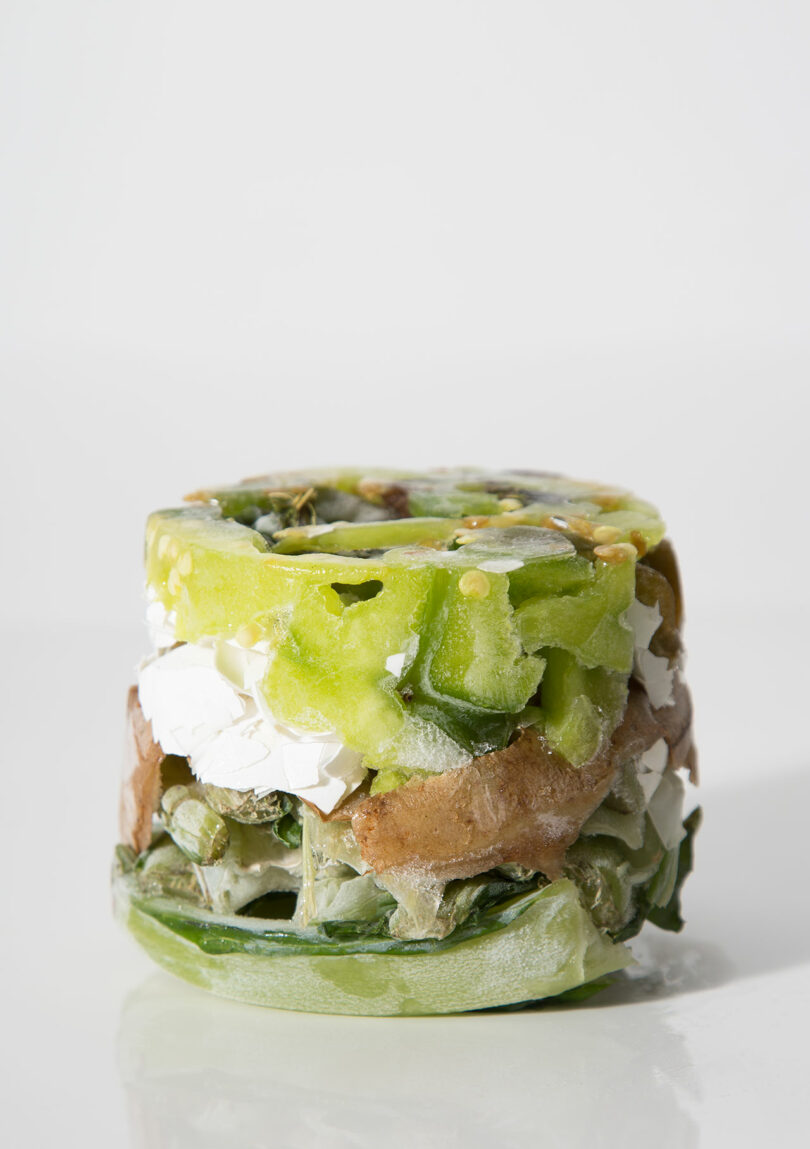 A frozen stack of mixed vegetables and white cheese sits on a white surface with a plain white background.