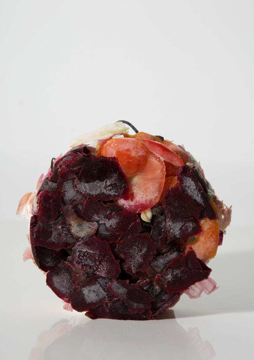 A tightly packed sphere made of assorted vegetable peels, including beets, onions, and carrots, set against a plain white background.