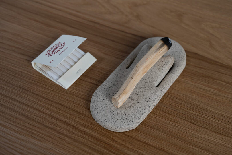 A bundle of white matches in a paper case and a charred wooden stick resting on a stone holder, all placed on a wood surface.