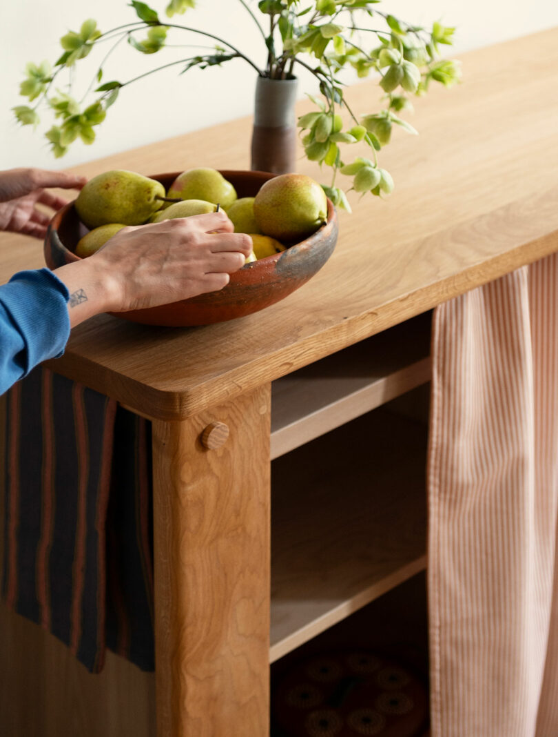A person places a bowl of pears on a wooden sideboard with striped towels hanging and a vase of flowers on top.