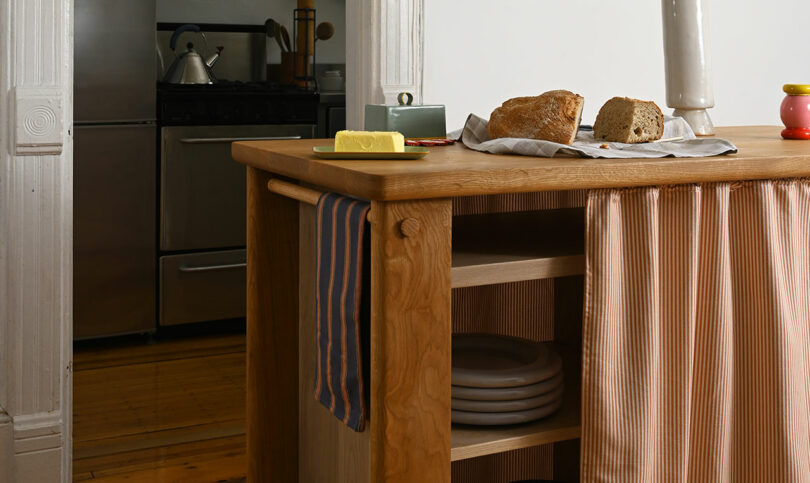 A wooden kitchen island with sliced bread, butter, and a knife on top; striped towel and plates are visible, with a stove and kettle in the background.