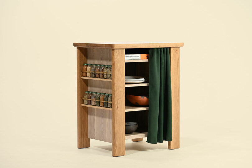 A small wooden kitchen island with open shelves holding plates, bowls, books, and jars, and a green curtain partially covering one side.