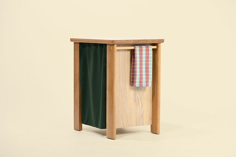 A wooden side table with a green fabric pocket on one side and a colorful checkered cloth draped over a horizontal bar.