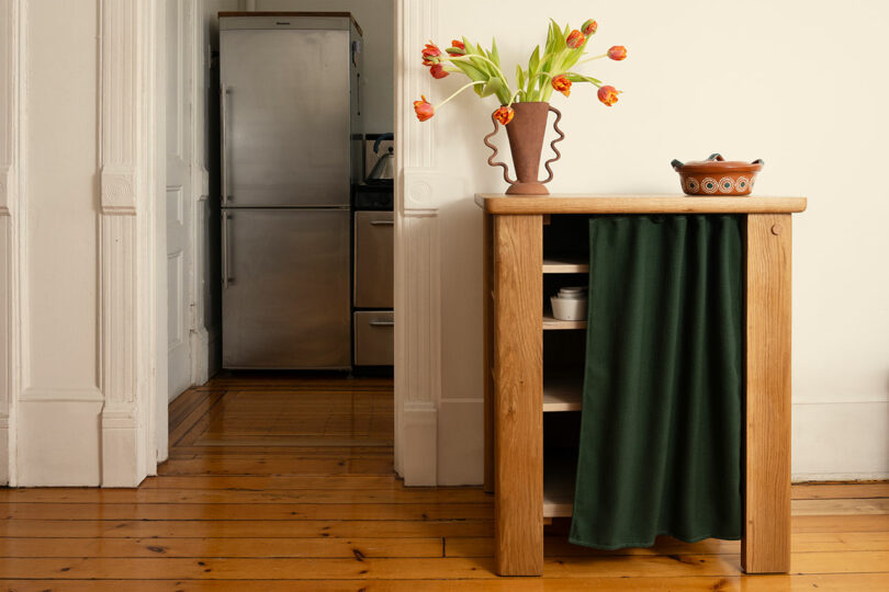 A wooden kitchen shelf with a green curtain holds a ceramic pot and vase of orange tulips; a stainless steel refrigerator is visible in the background.