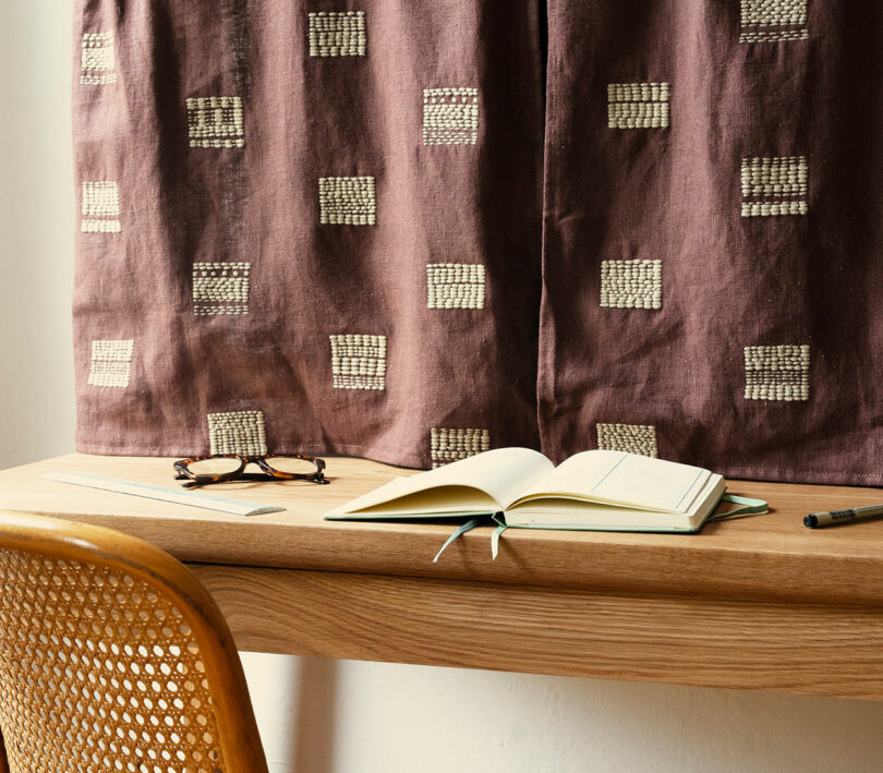 A wooden desk with an open notebook, pen, and eyeglasses sits in front of a window covered by patterned curtains; a woven chair is tucked under the desk.